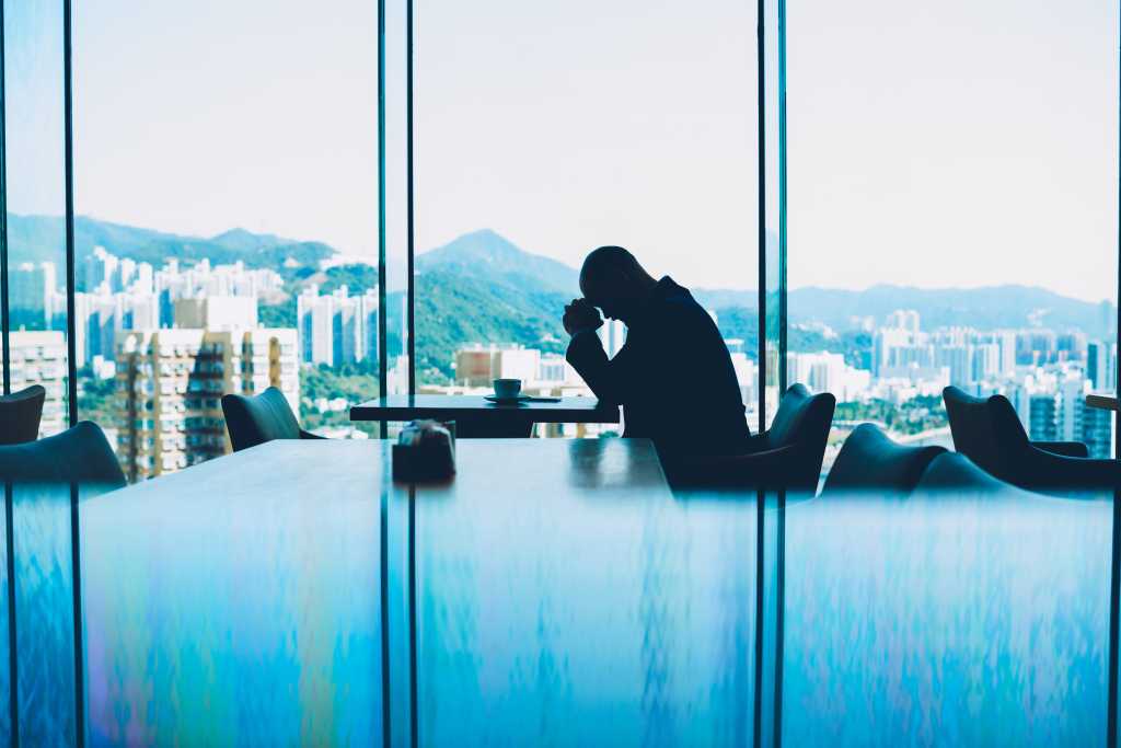 Silhouette of depressed male proud CEO tired from hard work and unsuccessful business project sitting at table with cup of coffee in modern interior. Alone entrepreneur in crisis hold head in despair