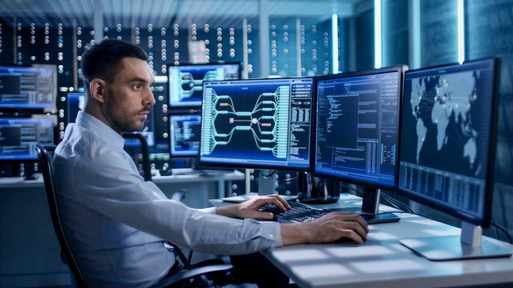 System Security Specialist Working at System Control Center. Room is Full of Screens Displaying Various Information.