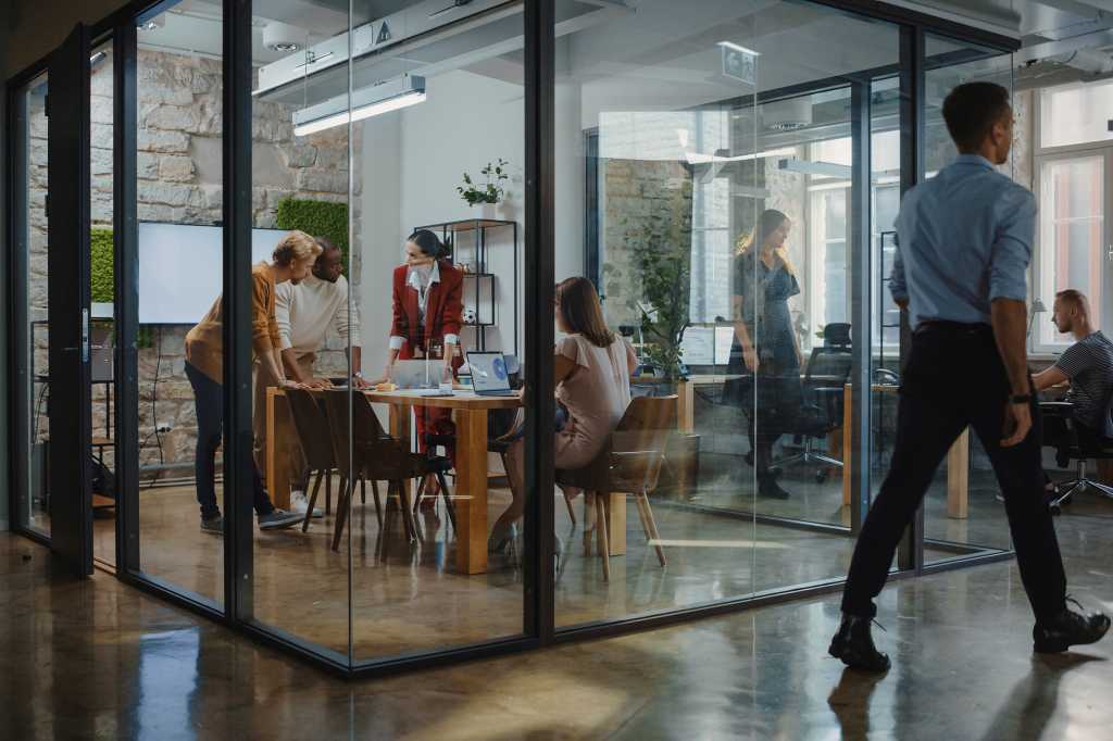 A group discussion takes place in a glass conference room, in a busy office workspace.