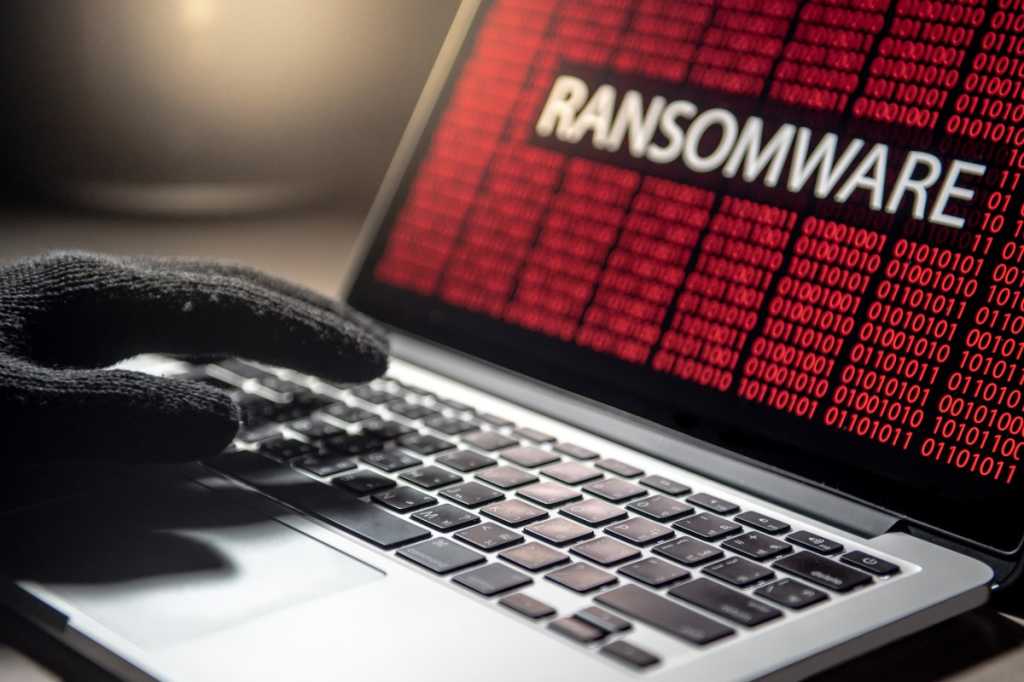 A photograph of a laptop showing binary in red text, with ransomware written in white in a black rectangle in the middle of the screen. A hand in a black glove can be seen resting on the keyboard of the laptop.