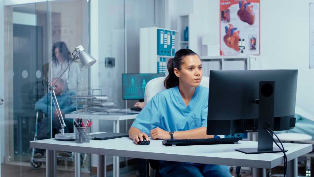 Nurse working on PC in modern private clinic with glass walls where patients are brought in wheelchair from an elevator and other persons are going through hallway. Team of doctors researching