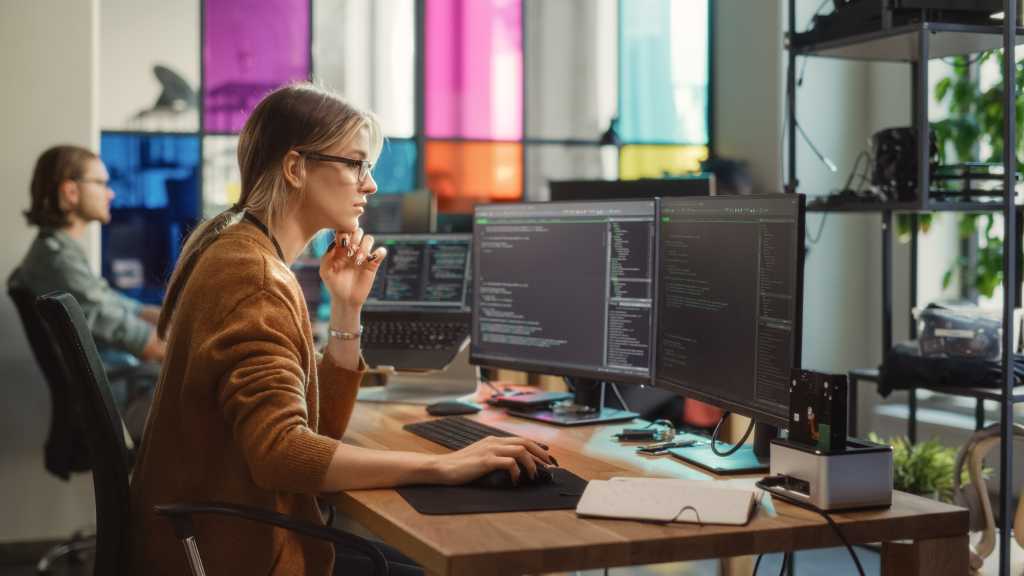 Caucasian Woman Writes Lines of Code on Desktop PC with Multiple Displays and a Laptop in Stylish Office. Professional Female Data Scientist Using Software To Analyze Information from Internet.