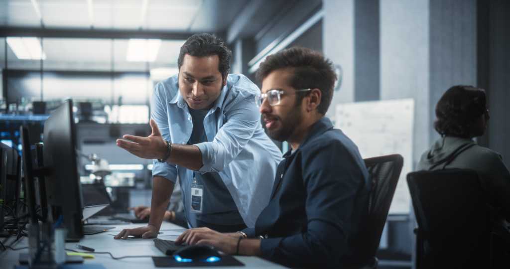 Creative Team of Indian Specialists Having a Conversation at a Workplace Next to Computer. Two Male Software Engineers Discussing a Software Code for Their Digital Blockchain Development Project