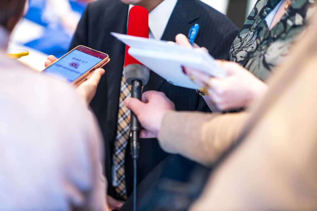 News conference, journalists taking notes, holding microphone and digital voice recorder interviewing unrecognizable politician or business person during media event