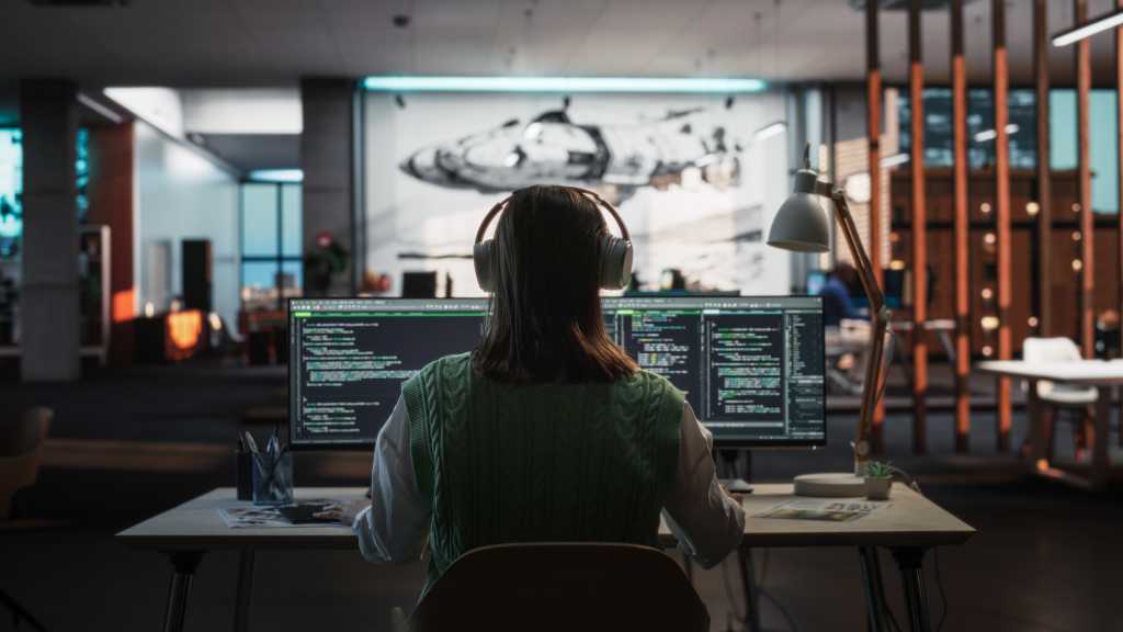 Creative Office: Professional Female Programmer Uses Headphones, Working on Desktop Computer. Focused Coder, Software Engineer Developing App, Video Game. Listening to Podcast or Music. Back View.