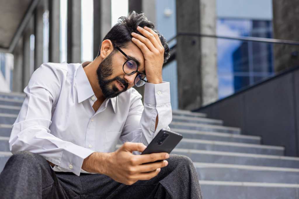 Young man depressed, sad dissatisfied and unhappy outside office building, holding phone, reading bad news from smartphone, businessman in shirt after work.