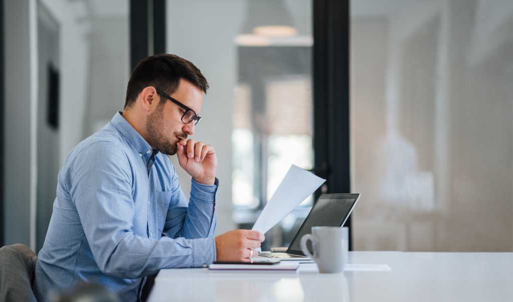 Serious pensive thoughtful young businessman or entrepreneur in modern contemporary office looking at and working with laptop and paper documents making serious and important business decision